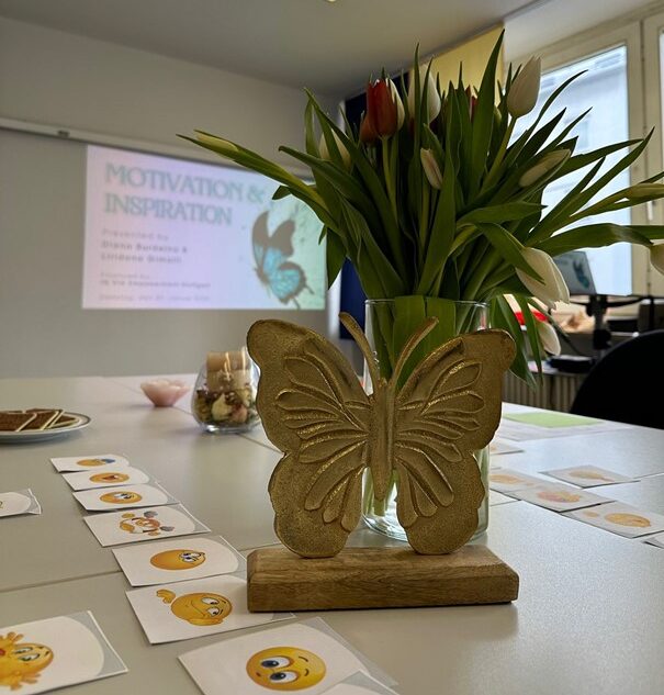 Ein Beamer wirft den Titel des Workshops "Motivation und Inspiration" an die Wand. Im Vordergrund stehen Blumen auf dem Tisch sowie Karten mit verschiedenen Emotions-Bildern